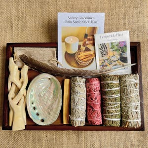Peut inclure: Un plateau en bois présente une collection d'articles spirituels. On y trouve des fagots de sauge, des bâtons de palo santo, une plume, une coquille d'ormeau et des livrets d'information. Le plateau est posé sur un fond texturé neutre.