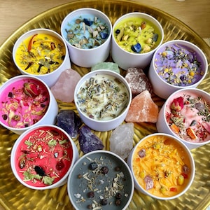 May include: A collection of colorful, scented candles in white containers, each adorned with dried herbs, flowers, and crystals. The candles are arranged on a gold tray, with various crystals scattered around them. The candles are in shades of yellow, blue, green, pink, red, and orange.
