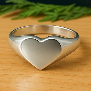 Puede incluir: Un anillo plateado con forma de corazón. El anillo se encuentra sobre una superficie de madera, con flores amarillas y follaje verde en el fondo. El corazón del anillo es liso y pulido.