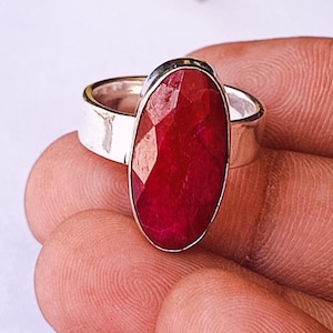 Könnte beinhalten: Ein Silberring mit einem großen, oval geschliffenen Rubin. Der Rubin hat eine tiefrote Farbe und ist in einem schlichten, polierten Silberband gefasst. Der Ring wird auf einer Hand gezeigt.