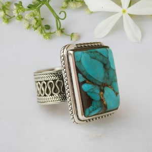 May include: A silver ring with a large rectangular turquoise stone. The turquoise has brown and gold veining. The ring band has a decorative pattern. The ring is set against a white background with flowers.