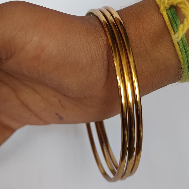 May include: Three gold-colored bangles are stacked on a wrist. The bangles are smooth and shiny, reflecting light. The wrist has a light skin tone. A green and yellow string bracelet is visible on the wrist.