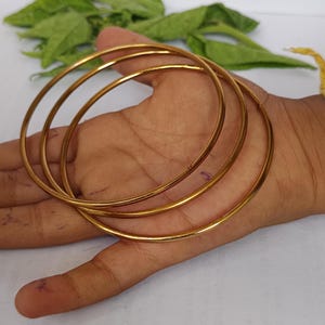 May include: Three gold-colored bangles are displayed, showcasing a simple, elegant design. The bangles are thin and circular, with a smooth, polished finish. The jewelry is presented on a light-toned background, highlighting the bangles' metallic sheen. The bangles are a classic jewelry item.