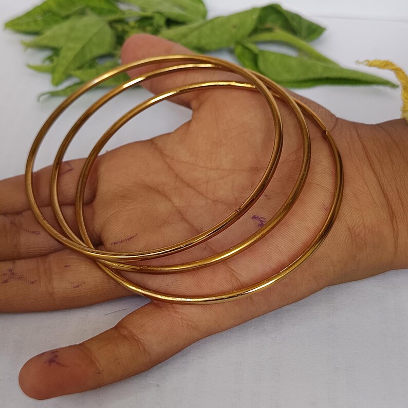 May include: Three gold-colored bangles are displayed, showcasing a simple, elegant design. The bangles are thin and circular, with a smooth, polished finish. The jewelry is presented on a light-toned background, highlighting the bangles' metallic sheen. The bangles are a classic jewelry item.