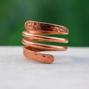 Puede incluir: Un anillo en espiral de color cobre con textura martillada. El anillo se enrolla alrededor del dedo varias veces, creando un diseño único. El metal tiene un tono marrón rojizo cálido. Una joya elegante.