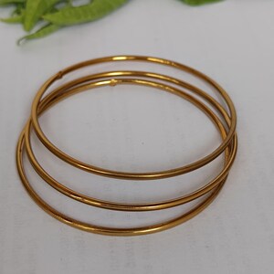 May include: Three gold-colored bangles are stacked on a white surface. The bangles are thin and circular, with a smooth, reflective surface. The jewelry is simple and elegant, suitable for everyday wear or special occasions.