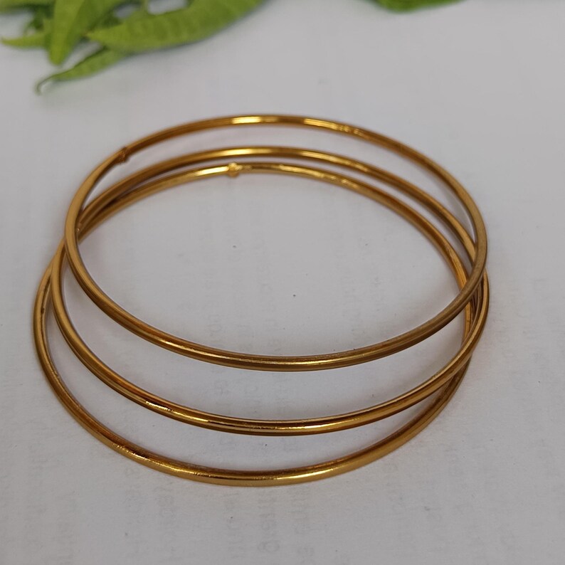 May include: Three gold-colored bangles are stacked on a white surface. The bangles are thin and circular, with a smooth, reflective surface. The jewelry is simple and elegant, suitable for everyday wear or special occasions.