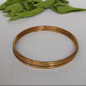 May include: A set of four gold-colored bangles, each with a smooth, rounded surface. The bangles are stacked together, creating a layered effect. Green leaves are visible in the background. The bangles are a classic jewelry item.