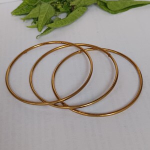May include: Three gold-colored bangles are arranged on a white surface, with green leaves in the background. The bangles are thin and circular, reflecting light. The jewelry is simple and elegant.