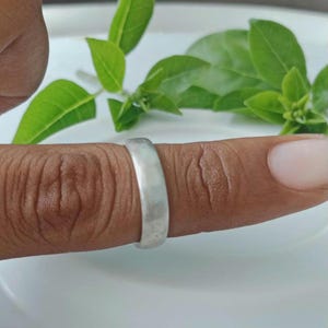 Könnte beinhalten: Ein silberner Ring mit gehämmertem Muster wird an einem Finger getragen. Der Ring hat ein schlichtes Banddesign. Grüne Blätter sind im Hintergrund, und der Finger hat einen hellrosa Nagel.
