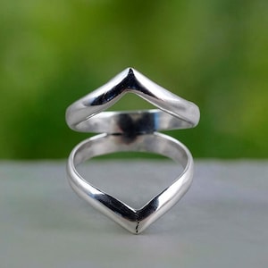 Op de afbeelding: Een zilveren ring met een V-vormig ontwerp. De ring heeft een gepolijst, reflecterend oppervlak en een minimalistische esthetiek. Het ontwerp bestaat uit twee V-vormen, de ene boven de andere gestapeld, waardoor een gelaagd effect ontstaat. De ring is waarschijnlijk gemaakt van sterling zilver.