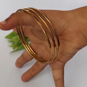 May include: A set of four gold-colored bangles. The bangles are thin, round, and smooth, reflecting light. The jewelry is displayed on a hand against a white background. The bangles are a classic accessory.