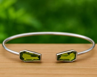 May include: A silver-coloured open cuff bracelet featuring two olive-green rectangular gemstones. The bracelet is presented on a wooden surface with a blurred green backdrop.