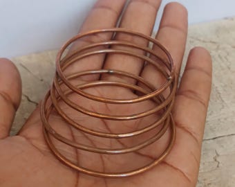 May include: A stack of six copper-colored metal bangles. The thin, round bracelets are stacked on top of each other, with a slight gap between each. The bracelets are resting on a person's open palm.