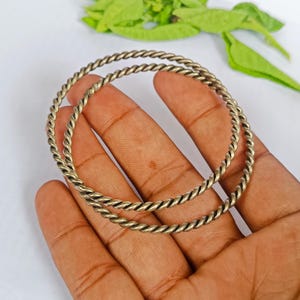 May include: Two antique brass bangles with a twisted rope design. The bangles are circular and appear to be made of a metal alloy. The bracelets are held in a hand against a white background with green leaves.