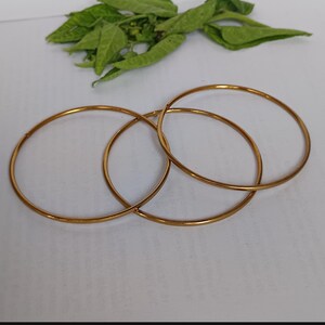 May include: Three gold-colored, thin, circular bangles are interlocked on a white surface. Green leaves are visible in the background. The bangles are simple in design and appear to be made of metal.