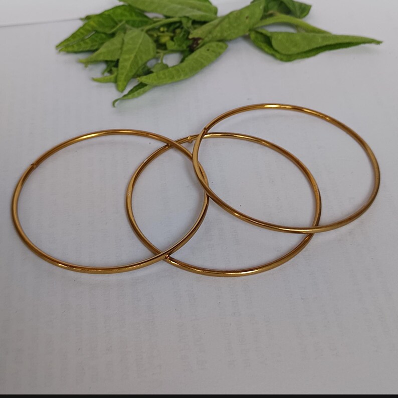May include: Three gold-colored, thin, circular bangles are interlocked on a white surface. Green leaves are visible in the background. The bangles are simple in design and appear to be made of metal.