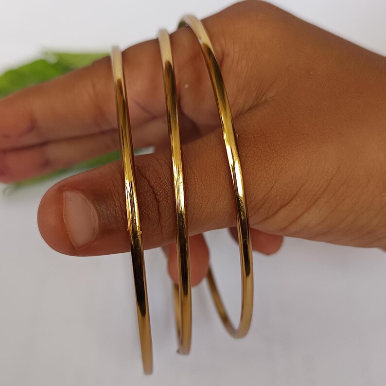 May include: Three gold-colored bangles are held between fingers. The bangles are thin and have a smooth, reflective surface. The jewelry is displayed against a neutral background.