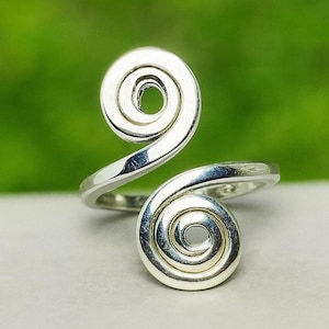 May include: A silver-colored ring with a spiral design. The ring features two swirling spirals, one at the top and one at the bottom, connected by a smooth, curved band. The ring is set against a green and white background.