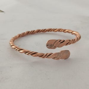 May include: A copper-colored open cuff bracelet with a twisted rope design. The bracelet has a simple, elegant style and is made of a shiny, reddish-brown metal. The ends of the bracelet are flattened and slightly curved.