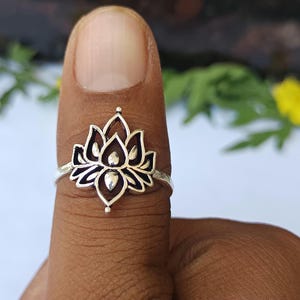 May include: A silver ring with a lotus flower design. The ring's floral centrepiece has detailed petals and a small, rounded centre. The ring is worn on a finger, showcasing its intricate design and polished finish.