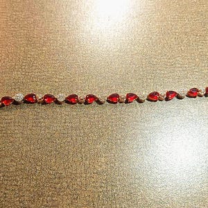 Peut inclure: Un bracelet en or orné de pierres précieuses rouges en forme de poire et de pierres rondes transparentes alternées. Le bracelet est posé sur une surface marron. Une étiquette blanche est attachée.