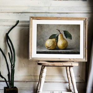 May include: A framed art print featuring a still life painting of two yellow pears with green leaves. The pears are positioned on a dark surface against a neutral, textured background. The artwork is enclosed in a light-colored wooden frame.