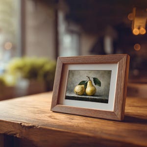 May include: A rectangular wooden picture frame containing a print of two yellow pears with green leaves. The frame has a light brown wood grain finish and a white mat border surrounding the central artwork, which depicts the fruit on a dark surface.