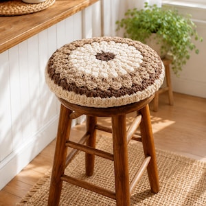 May include: A round, crocheted stool cover in shades of brown, beige, and cream sits atop a wooden stool. The cover features a circular pattern with a dark brown center and lighter concentric rings. The stool has a classic, three-legged design.