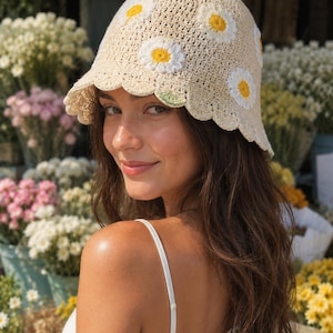 May include: A cream-colored crochet bucket hat adorned with white and yellow daisy appliqués. The hat features a scalloped brim and a textured, open weave design. The hat is a stylish accessory.