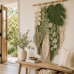 May include: A macrame wall hanging with crochet leaf designs in green and cream hues. The leaves are suspended from a wooden dowel with hanging cords. The wall art is displayed above a wooden bench, with a vase and a woven basket adding to the decor.