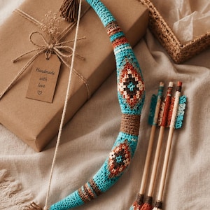 May include: A handmade crochet bow and arrows set. The bow is turquoise with brown and orange geometric patterns. Three arrows with wooden shafts and crochet fletching are included. A gift box with a tag that says "Handmade with love" is in the background.