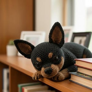 May include: A handmade crochet chihuahua dog in black and tan. The dog is lying on a wooden shelf, with its head and paws hanging over the edge. The dog has large, dark eyes and a detailed snout. Books are stacked on the shelf.