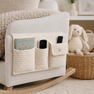 May include: A cream-coloured, crocheted chair organiser with three pockets attached to the side of a rocking chair. The pockets hold a book, a phone, and a remote control. The book is titled "Guess How Much I Love You".