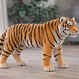 May include: A handcrafted crochet tiger figurine. The tiger has orange and black stripes, white accents, and a detailed face. The tiger is standing on four legs, with its tail extended. The figurine is approximately 24 inches long.