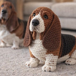 May include: Crocheted Basset Hound dog with brown, black, and white details. The handmade plushie has long floppy ears and a realistic appearance. Another dog is slightly out of focus in the background.