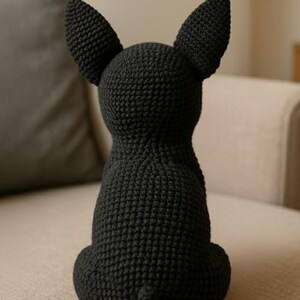 May include: A black crocheted dog figurine, viewed from the back, with large, upright ears and a small tail. The handmade dog is sitting on a beige sofa. The dog is made of a textured yarn.