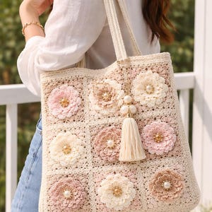May include: A cream-colored crochet tote bag with a floral design. The bag features nine squares, each adorned with a different colored flower, including pink, cream, and brown. A tassel and pearl accents hang from the top.