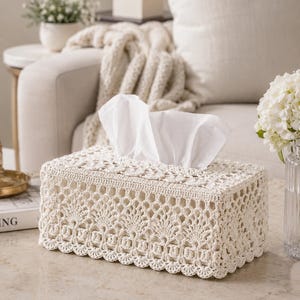 May include: Cream-colored rectangular tissue box cover with an intricate crochet design. White tissues are visible at the top. The cover is placed on a light-colored surface, with a neutral-toned background.