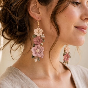 May include: Long, dangling earrings featuring a floral design. The earrings are made of crocheted flowers in shades of pink, mauve, and cream, with green and cream leaves. The earrings are suspended from gold-colored hooks.