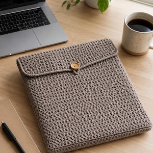May include: A crocheted laptop sleeve in a neutral brown color. The sleeve has a textured pattern and a flap closure secured with a wooden button. The sleeve is on a wooden desk next to a laptop, notebook, pen, and a mug of coffee.