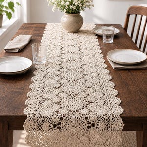 May include: A beige, crocheted table runner with a scalloped edge, placed on a dark wooden table. The runner features a detailed floral pattern. The table is set with plates, glasses, and a vase of white flowers.
