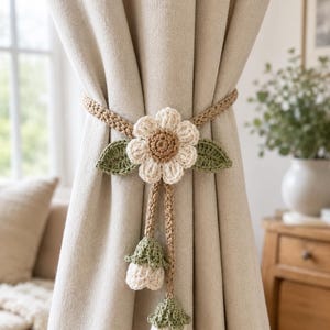 May include: Crocheted curtain tieback featuring a large flower with a beige center and white petals, flanked by green leaves. The tieback is beige and has two dangling flower buds. The curtains are a neutral color.