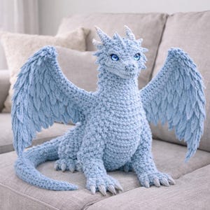Puede incluir: Un dragón de ganchillo azul claro con grandes alas, garras afiladas y una larga cola. El dragón tiene detalles, incluyendo cuernos, escamas y ojos azul brillante. Está sentado en un sofá gris.