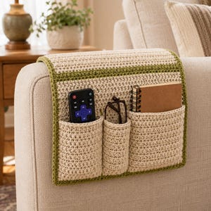 May include: Beige crocheted sofa armrest tidy with green edging. It has three pockets containing a remote control, spectacles, and a small notebook. Designed to fit over a sofa arm.