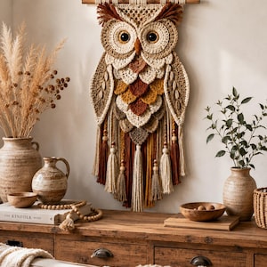 May include: A large, handmade owl wall hanging. The owl is crafted from beige, brown, and orange yarn, with large, dark eyes. It features a textured design with tassels and wooden beads. The owl is suspended from a wooden dowel, adding a rustic touch to the decor.