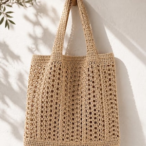 Può includere: Una borsa tote all'uncinetto beige a trama aperta con due manici lunghi, appesa a una parete bianca. Il corpo rettangolare e i manici della borsa sono realizzati con un filato spesso e strutturato. La borsa è sospesa a un gancio di legno.