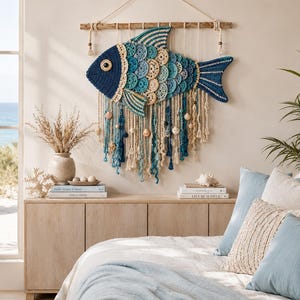 Op de afbeelding: Een grote, handgemaakte gehaakte vis wandhanger in tinten blauw en crème. De vis heeft een gedetailleerd schubbenpatroon en is opgehangen aan een houten staaf in drijfhoutstijl. Decoratieve kwasten en schelpen hangen eronder.
