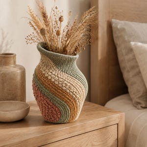 Puede incluir: Un jarrón de ganchillo multicolor hecho a mano con un diseño en espiral, lleno de hierbas ornamentales secas. El jarrón presenta rayas rosas, verdes, beiges y amarillas. El jarrón está sobre una mesita de noche de madera.