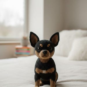 May include: A crocheted chihuahua dog figurine in black and tan. The handmade dog has large, upright ears and is sitting on a white surface. The dog's body is black with tan markings.